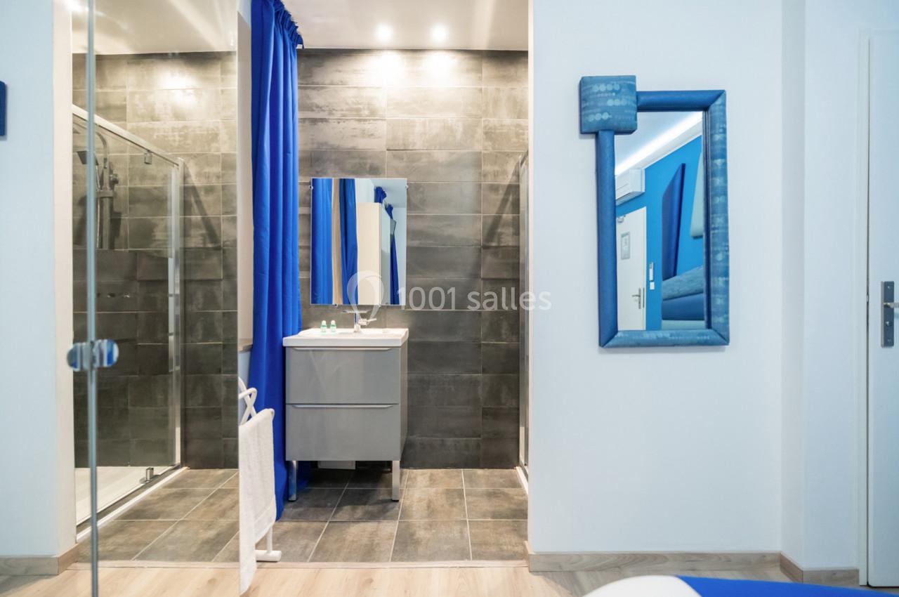 Salle de bain moderne avec douche vitrée, carrelage gris foncé, meuble lavabo et miroir encadré bleu.