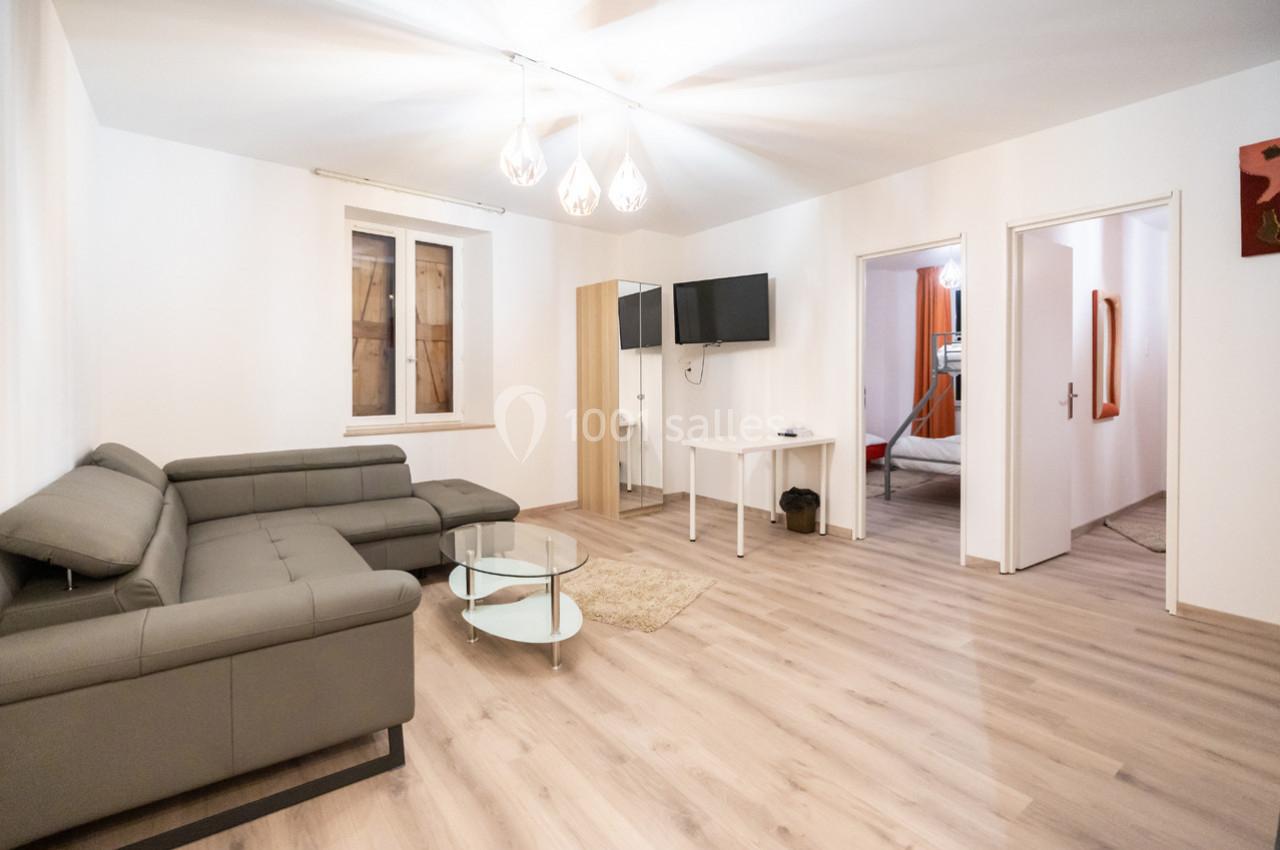 Salon lumineux avec canapé d'angle gris, table basse en verre, parquet clair et accès à une chambre adjacente.