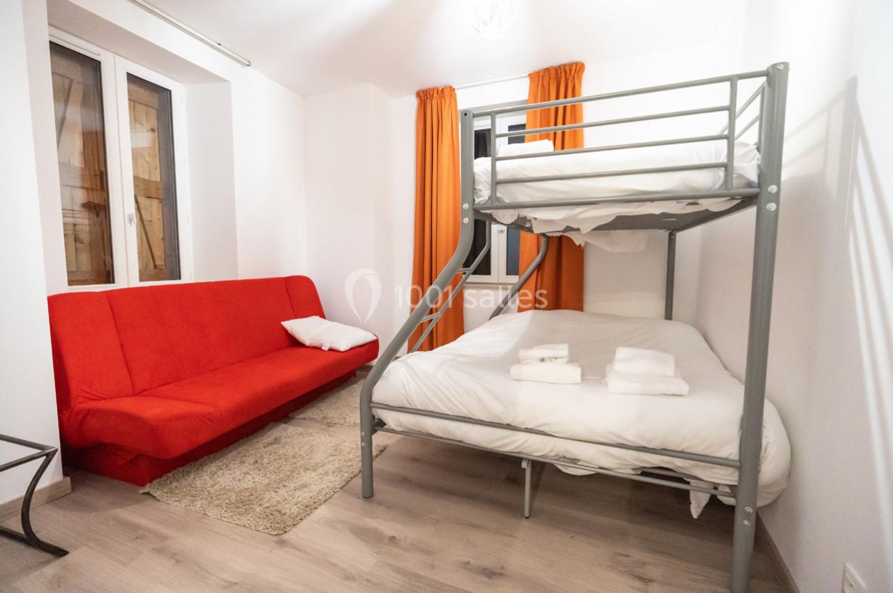 Chambre avec un lit superposé, un canapé-lit rouge, des rideaux orange et un sol en parquet clair.