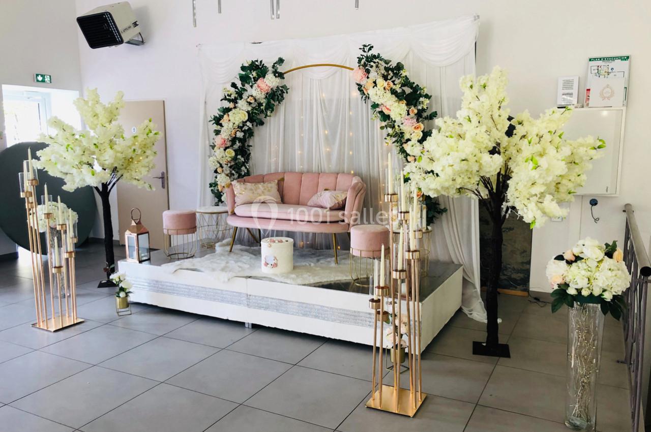 Décoration de salle avec arche florale, canapé rose, fleurs blanches et dorures sur une estrade.