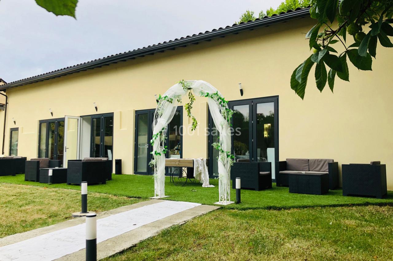 Allée menant à une arche décorée de voilages et de feuillages devant un bâtiment moderne avec terrasse aménagée.