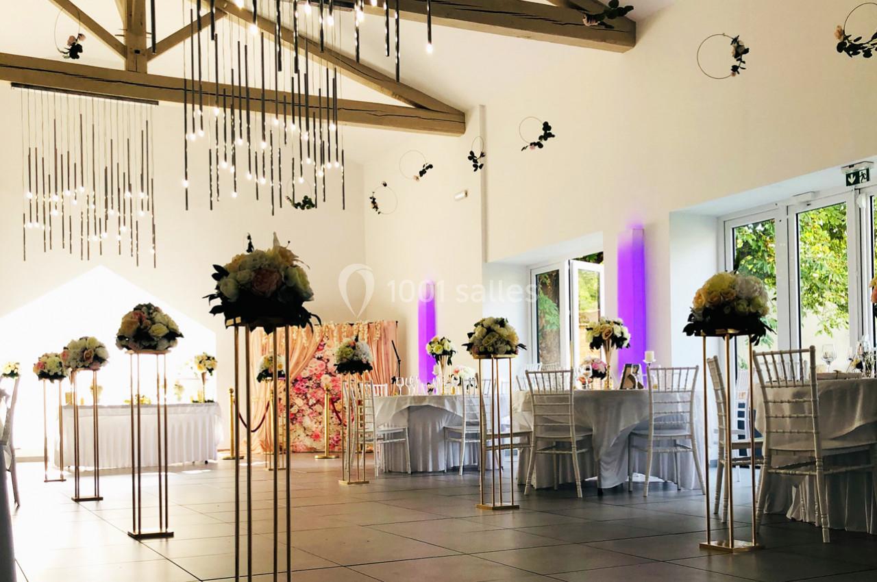 Salle de réception décorée avec des tables dressées, des chaises blanches et des arrangements floraux sur des supports dorés.