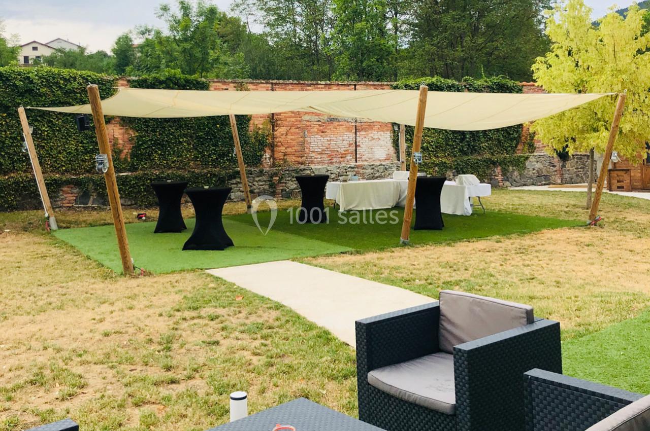 Espace extérieur aménagé avec tables hautes, chaises et voiles d'ombrage, entouré de verdure et d'un mur en briques.