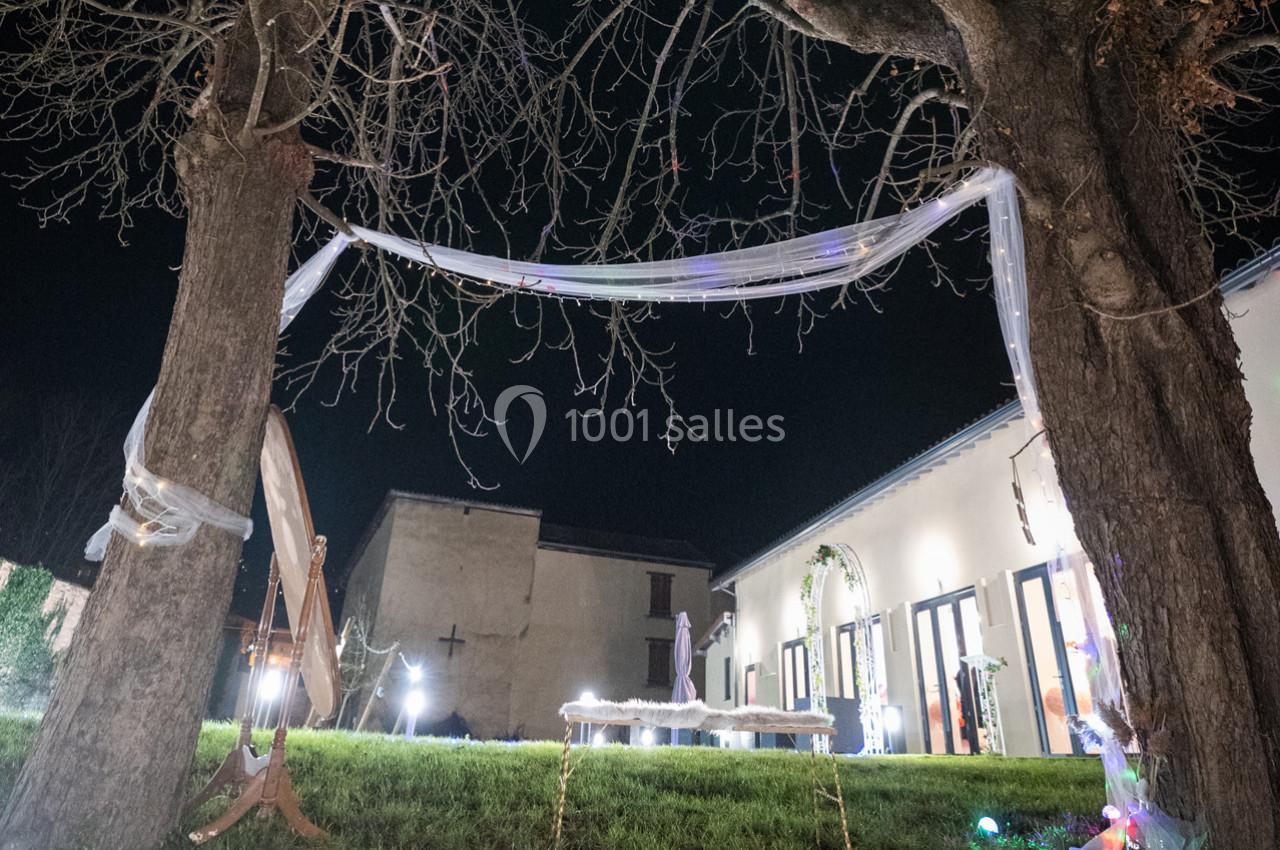 Décoration nocturne avec guirlandes lumineuses et voilages suspendus entre deux arbres, devant des bâtiments éclairés.