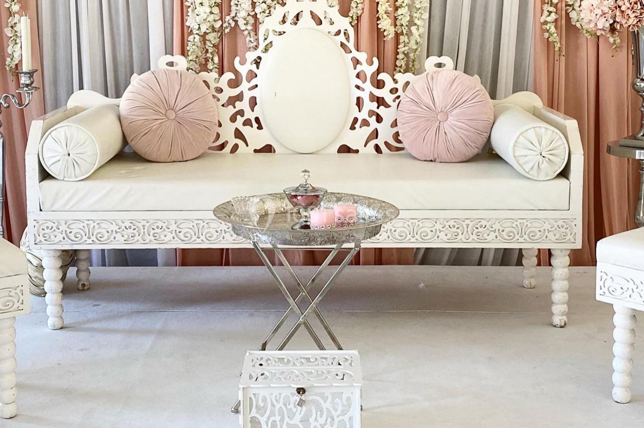 Canapé blanc sculpté avec coussins roses, table basse argentée et décor floral en arrière-plan.