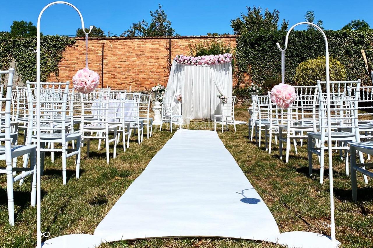 Allée centrale décorée pour une cérémonie en extérieur, bordée de chaises blanches et ornée de fleurs roses.