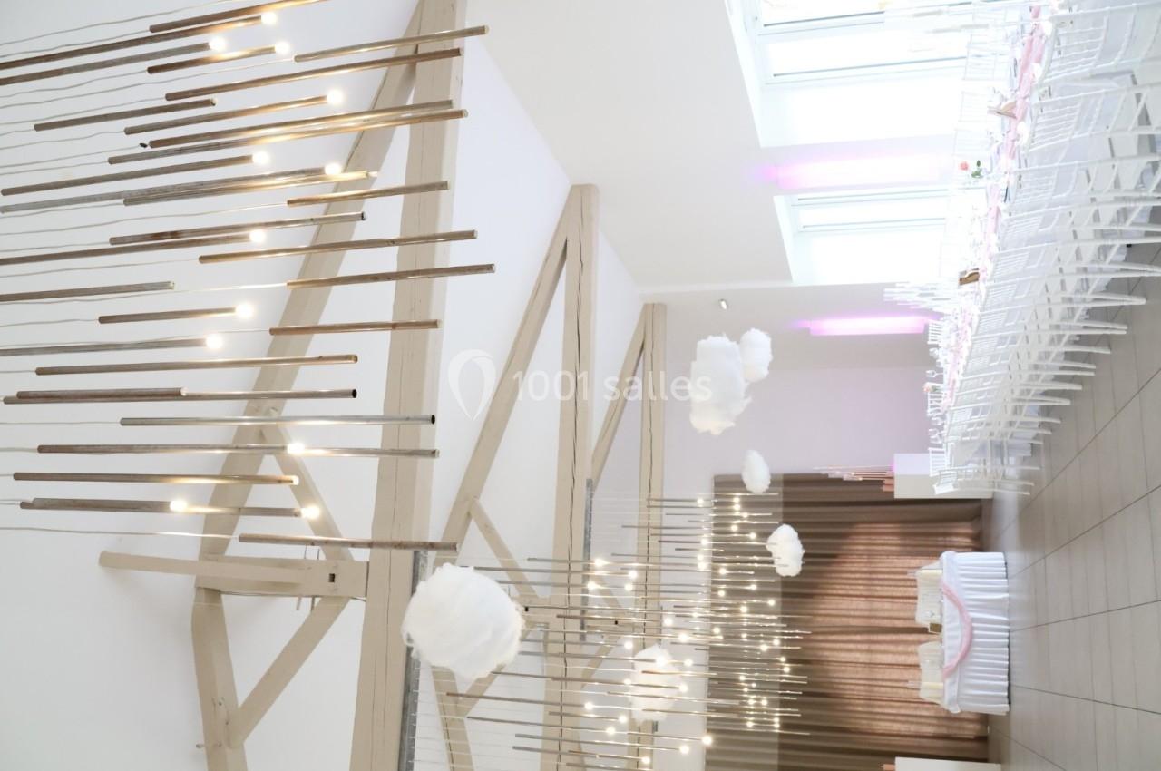 Salle de réception lumineuse avec des décorations suspendues, des tables dressées et des chaises blanches alignées.