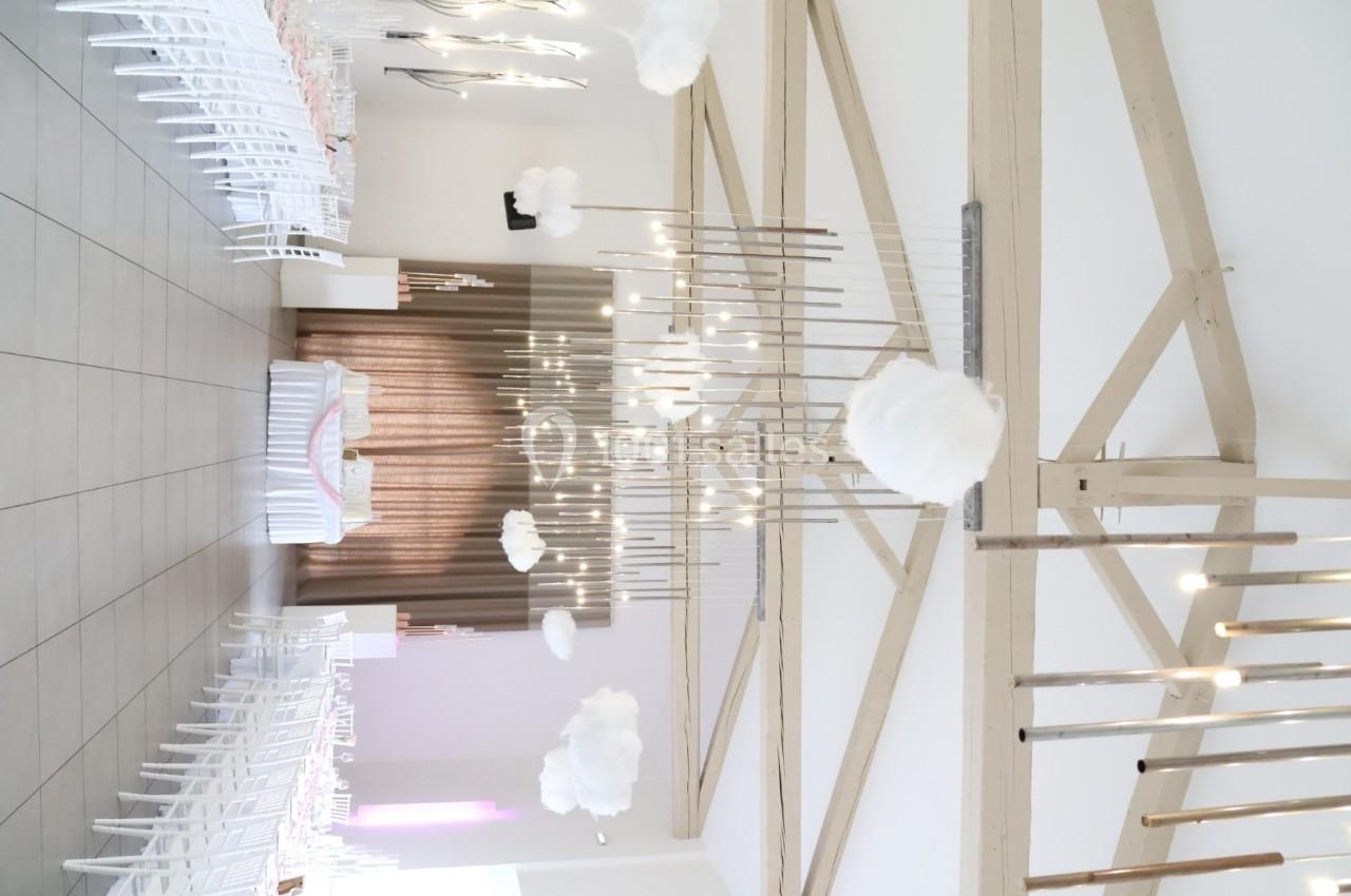 Salle décorée pour un événement avec des chaises blanches, des nuages suspendus et des lumières verticales.