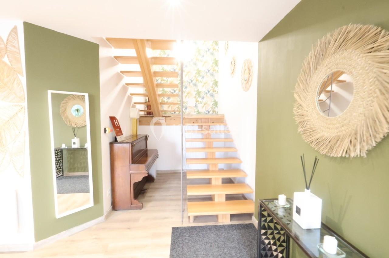 Escalier en bois dans un intérieur lumineux avec murs verts, miroir, piano et décoration murale en rotin.