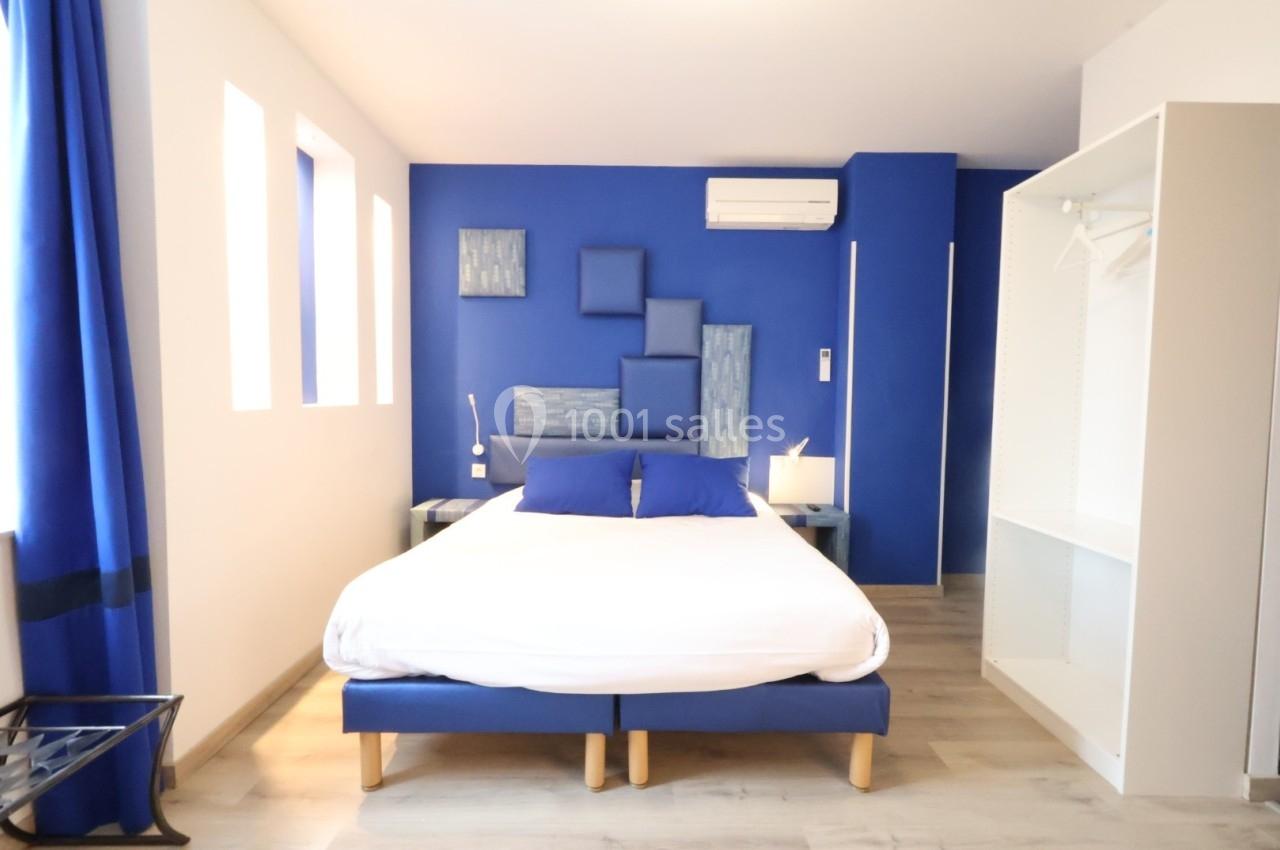 Chambre moderne avec murs bleus, lit double, éclairage naturel, armoire ouverte et climatisation.