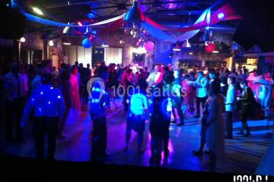 Salle de réception décorée avec des tables rondes, nappes blanches, chaises ornées de nœuds beige et éclairage vert.