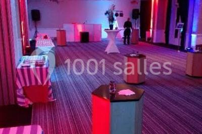 Salle de réception décorée avec des tables rondes, nappes blanches, chaises ornées de nœuds beige et éclairage vert.