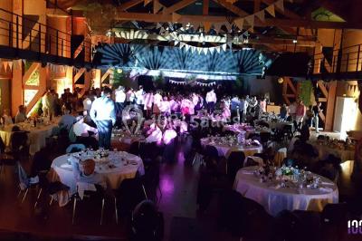 Salle de réception décorée avec des tables rondes, nappes blanches, chaises ornées de nœuds beige et éclairage vert.