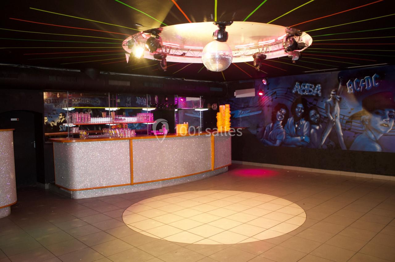 Salle de danse avec piste circulaire éclairée, bar scintillant et fresque murale colorée sous des lumières tamisées.