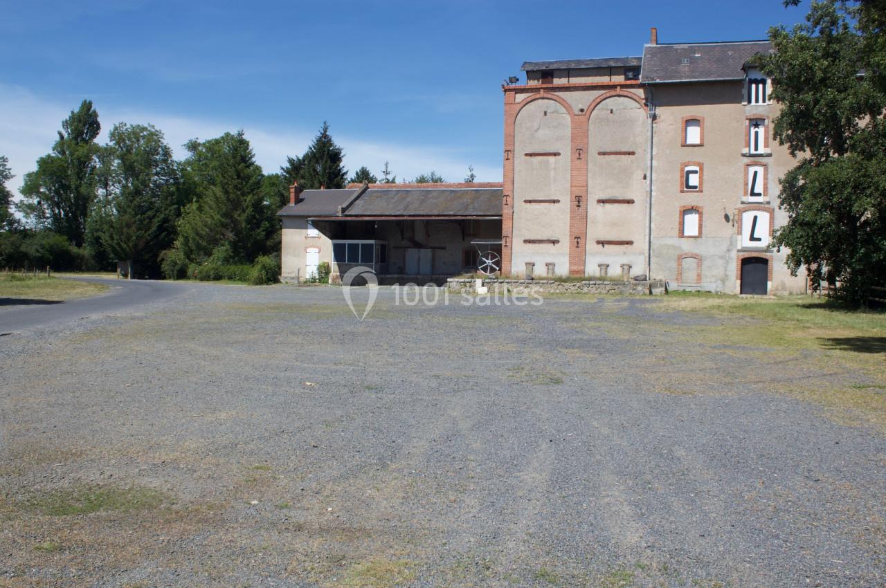Bâtiment ancien en briques et pierre, entouré d'arbres, avec une cour en gravier sous un ciel dégagé.