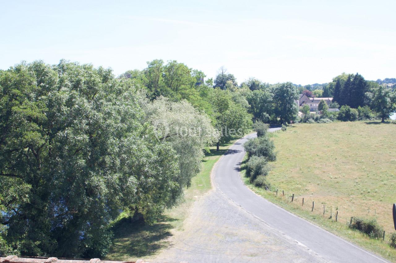 Route de campagne bordée d'arbres et de prairies, menant à un petit village sous un ciel dégagé.