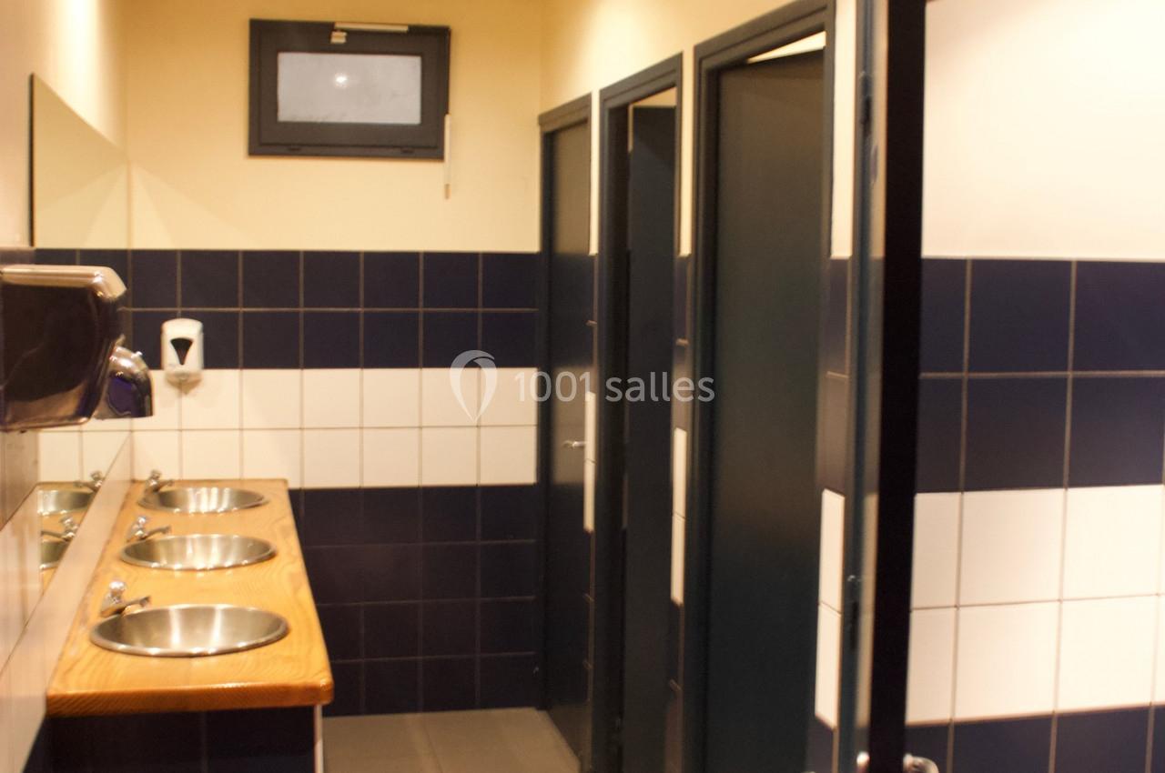 Salle de bain publique avec lavabos en inox, carrelage bleu et blanc, et portes de cabines fermées.