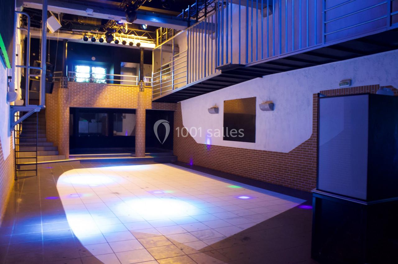 Salle vide avec piste de danse éclairée, murs en briques et métal, et mezzanine au fond.