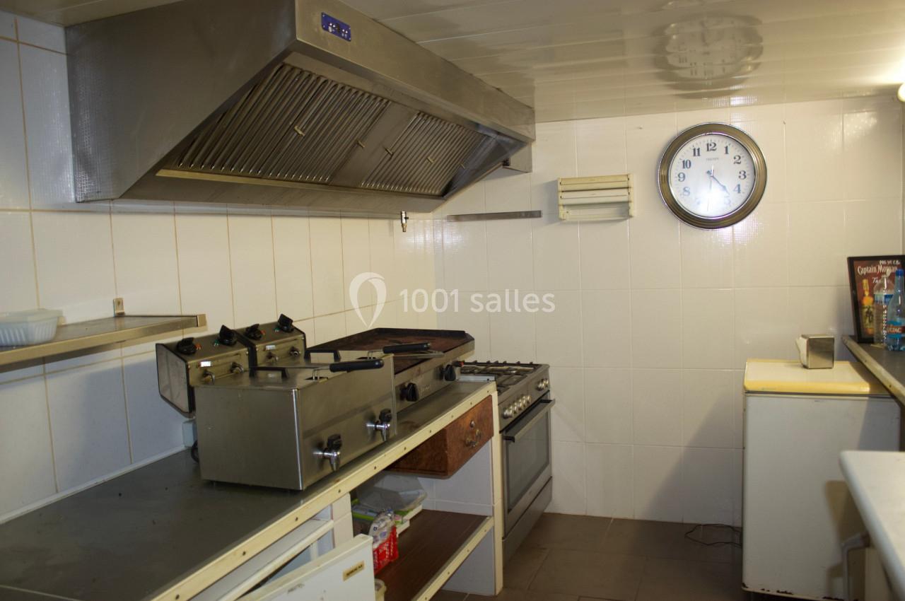 Cuisine professionnelle avec hotte, friteuse, four, plaques de cuisson et horloge murale sur un mur carrelé blanc.