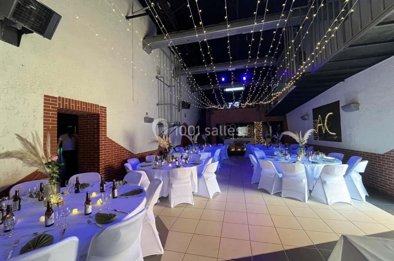 Salle décorée pour un événement avec tables rondes, nappes blanches, guirlandes lumineuses suspendues et éclairage tamisé.