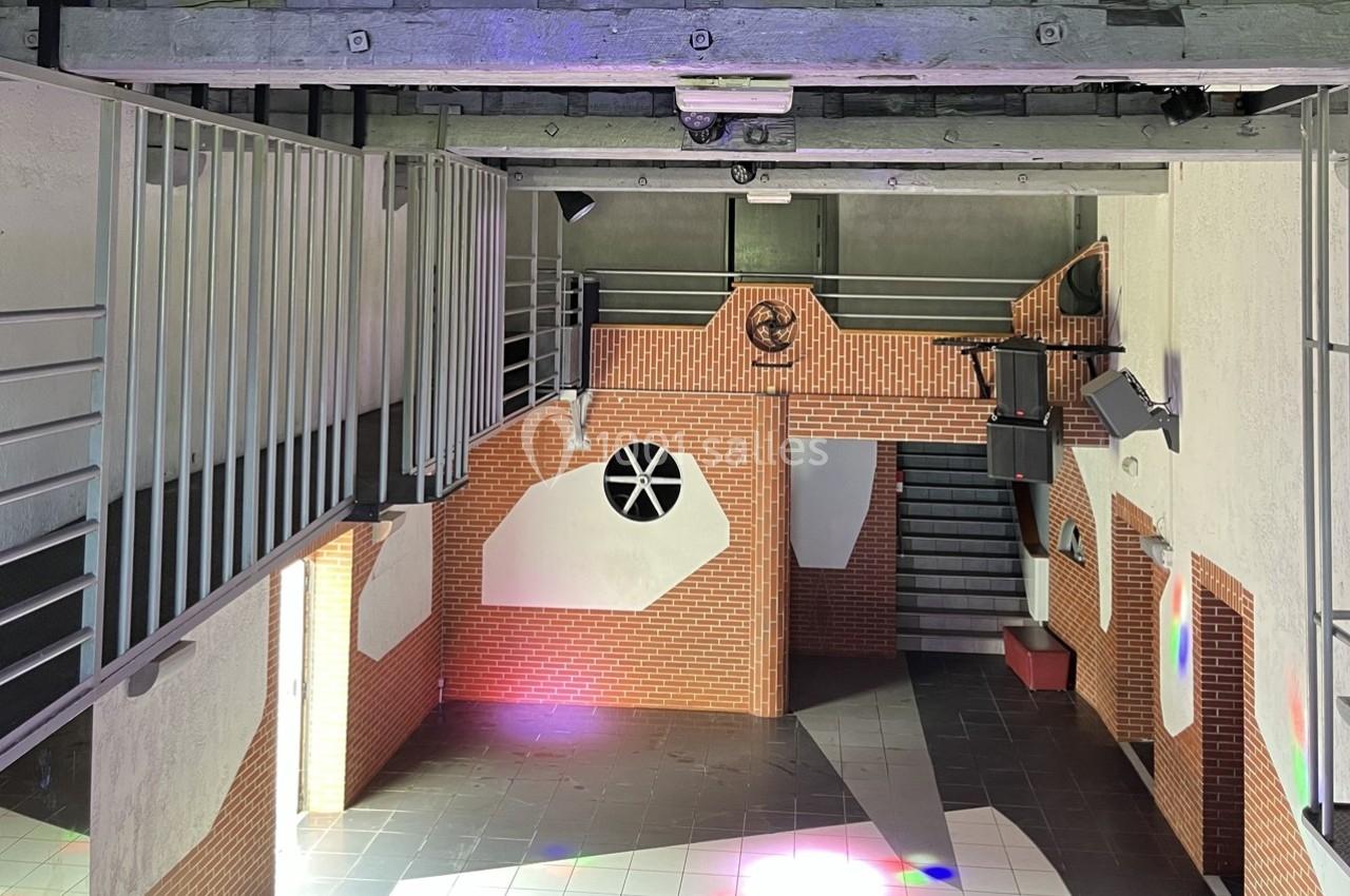 Intérieur d'un bâtiment avec murs en briques, mezzanine métallique, escalier et jeux de lumière colorés au sol.