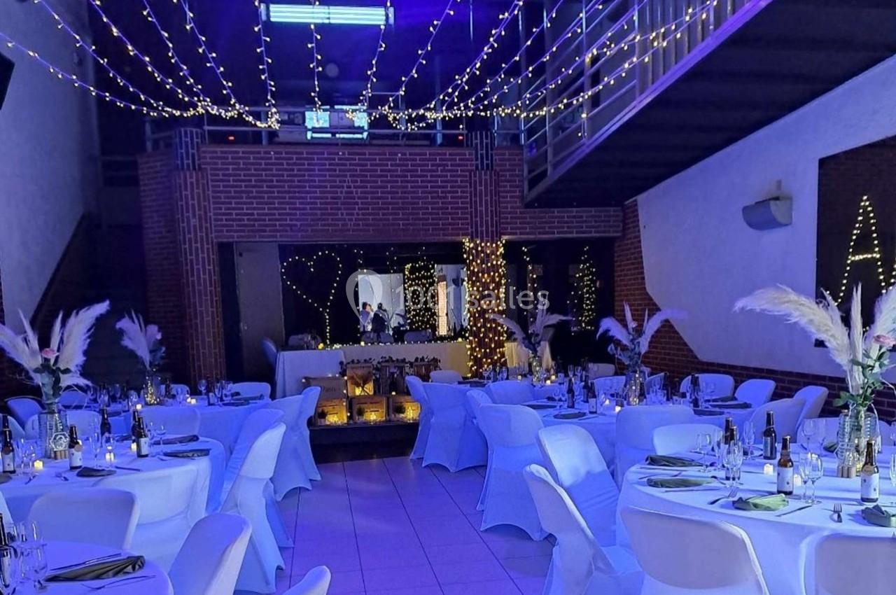 Salle décorée pour un événement, avec tables dressées, guirlandes lumineuses suspendues et ambiance tamisée.