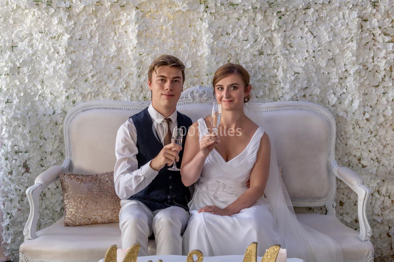 Un couple assis sur un canapé blanc, tenant des flûtes de champagne, devant un mur décoré de fleurs blanches.