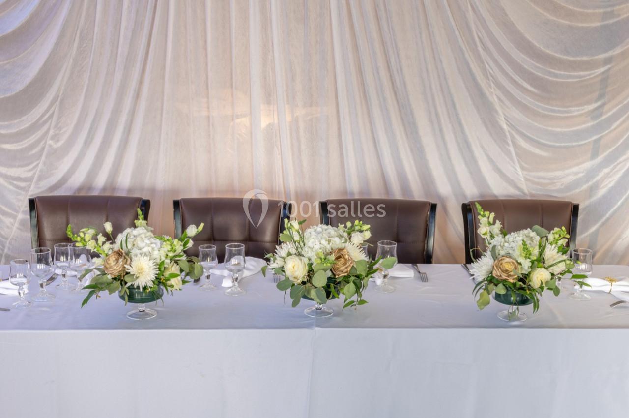 Table décorée avec nappes blanches, chaises en cuir marron et arrangements floraux élégants devant un rideau drapé.