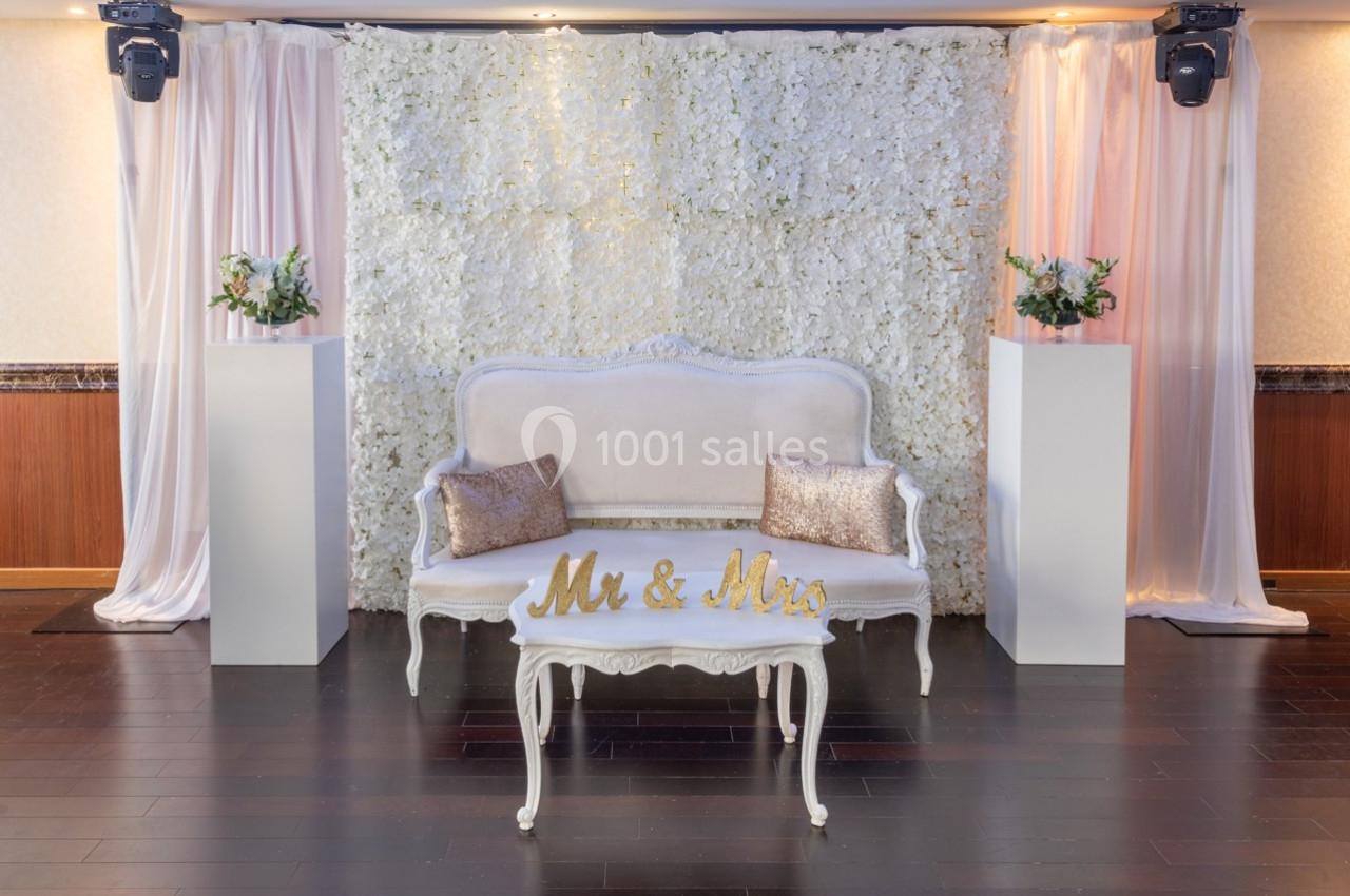 Banc blanc orné de coussins dorés devant un mur de fleurs blanches, encadré de rideaux et de colonnes décoratives.