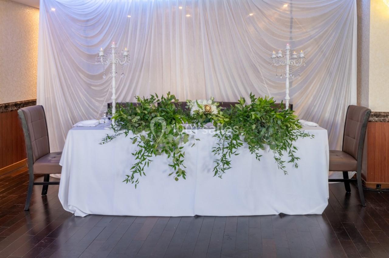 Table décorée avec une nappe blanche, ornée de feuillage, chandeliers et fleurs, entourée de deux chaises en cuir brun.