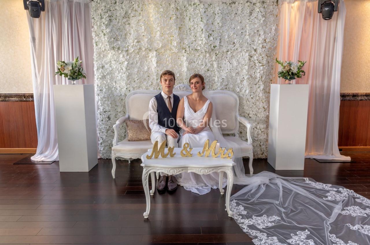 Un couple de mariés assis sur un canapé devant un mur fleuri, avec une table portant l'inscription ’Mr & Mrs’.