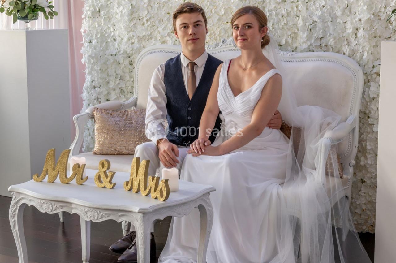 Un couple en tenue de mariage est assis sur un canapé devant un mur de fleurs blanches, avec une décoration ’Mr & Mrs’.