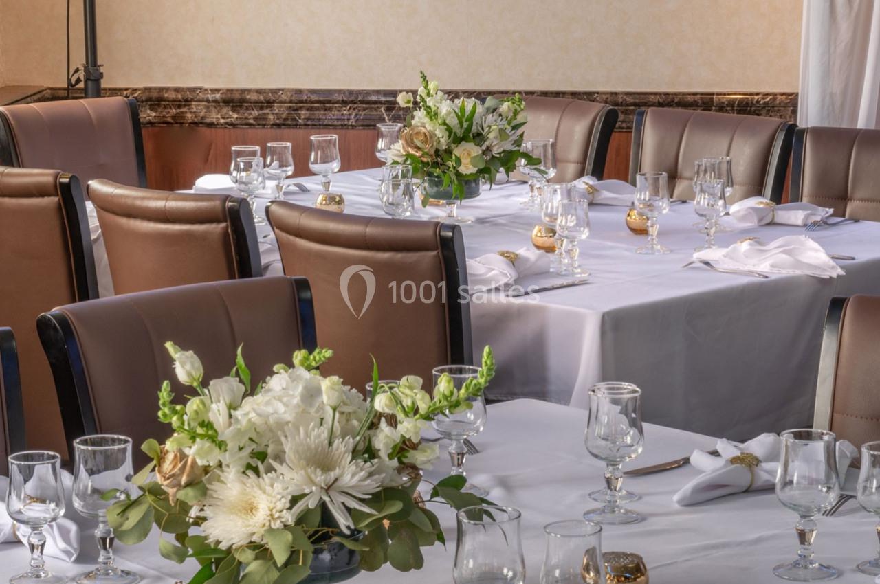 Salle de réception avec tables dressées, nappes blanches, verres, serviettes pliées et bouquets de fleurs.
