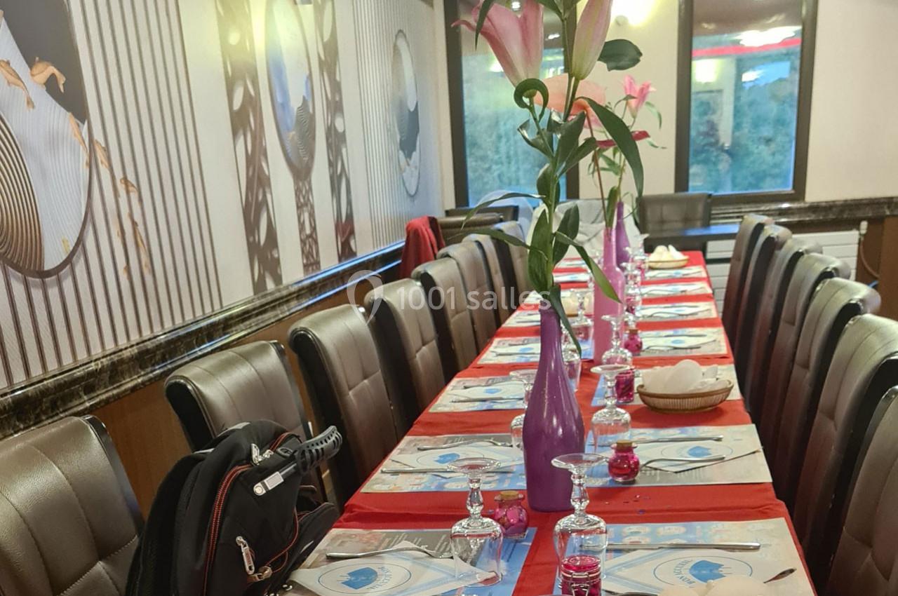 Table dressée dans un restaurant avec nappes rouges, verres alignés, vases violets contenant des fleurs roses.