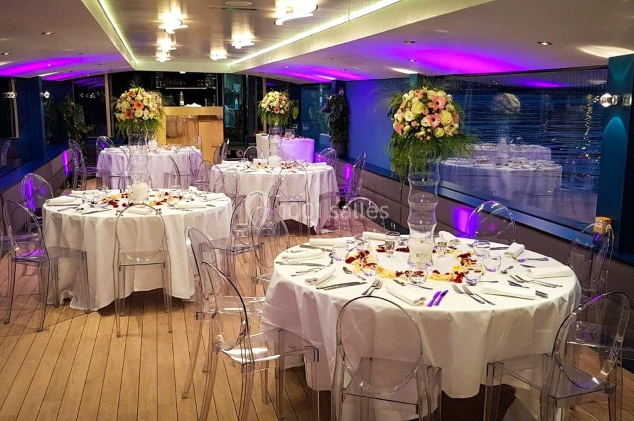 Salle de réception élégante sur un bateau, avec tables rondes dressées, chaises transparentes et décor floral.