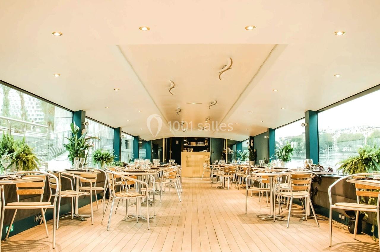 Salle lumineuse avec grandes baies vitrées, tables et chaises en métal, décorée de plantes vertes.