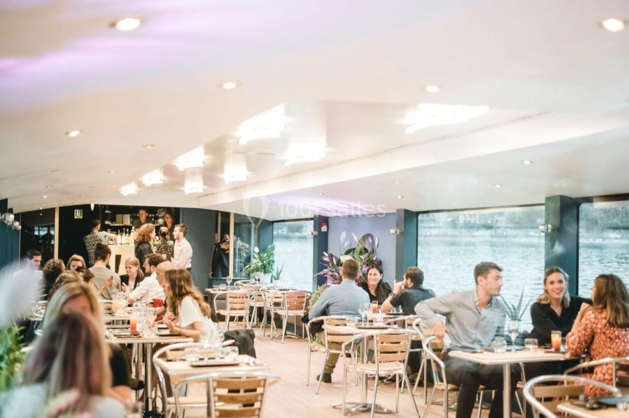 Salle lumineuse avec des tables et des chaises, des convives discutant et un bar en arrière-plan près d'une baie vitrée.