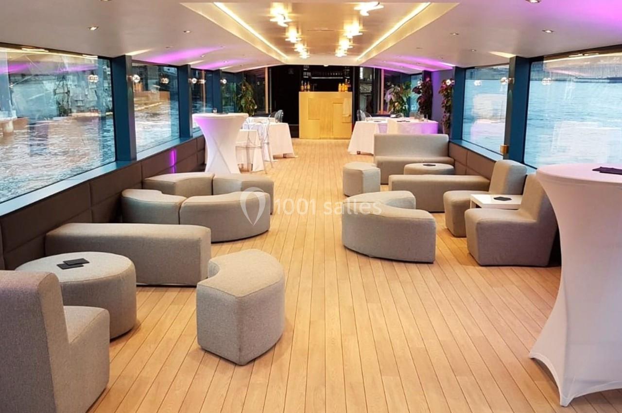 Intérieur d'un bateau avec salon moderne, mobilier gris, tables hautes et vue sur l'eau à travers de grandes baies vitrées.