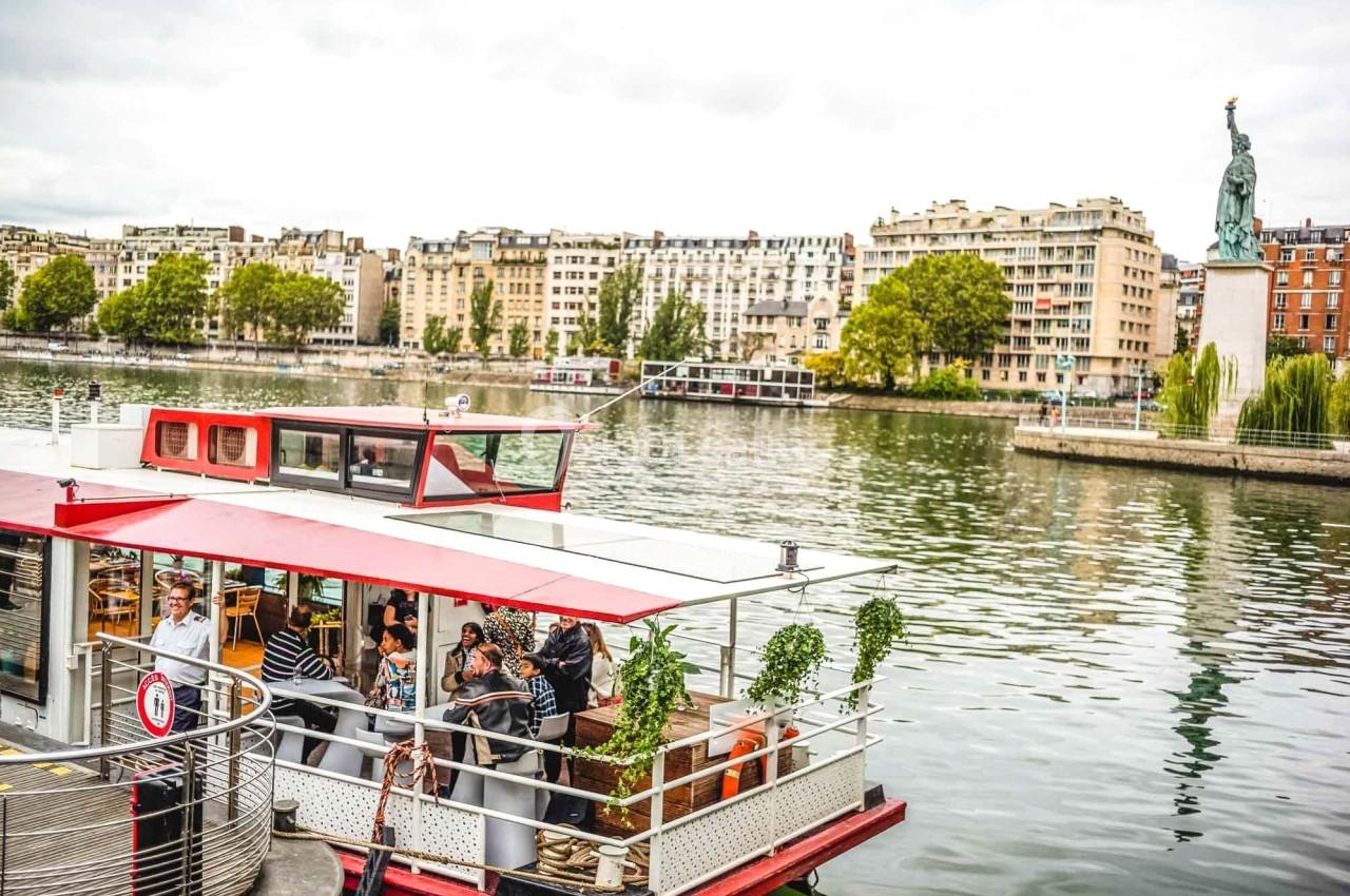 Bateau avec des passagers amarré sur une rivière, immeubles et réplique de la Statue de la Liberté en arrière-plan.