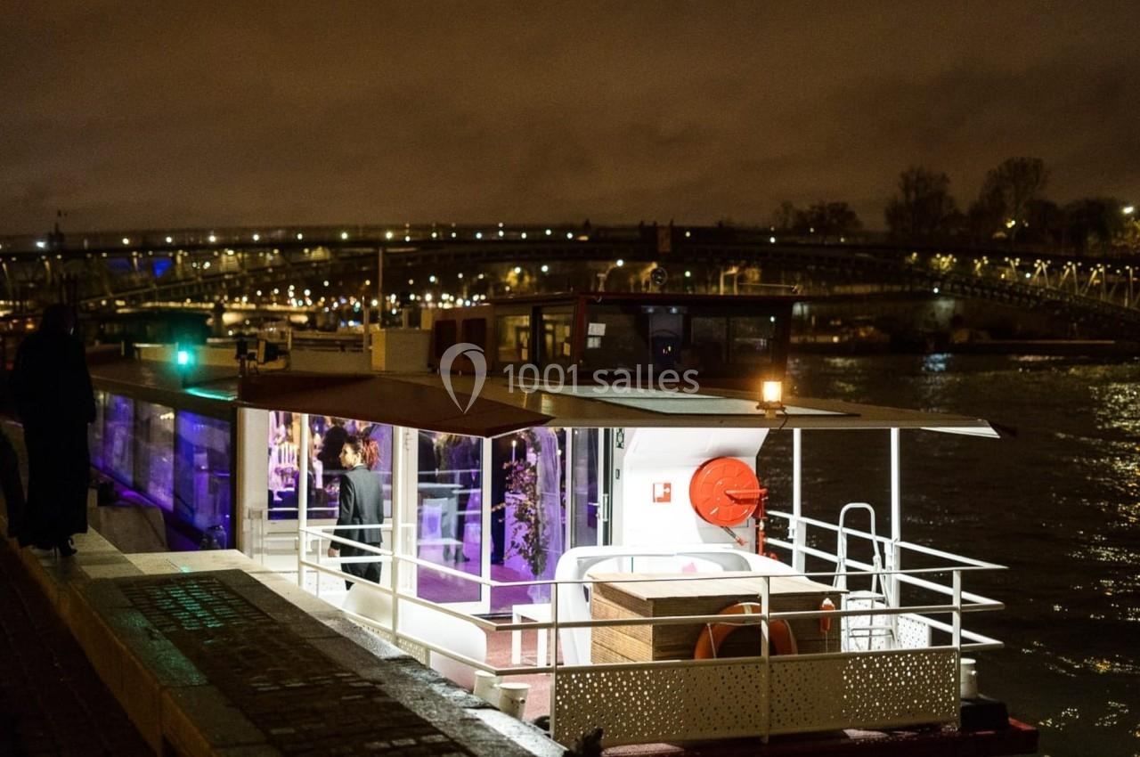 Bateau amarré au bord d'une rivière la nuit, éclairé par des lumières et avec un pont visible en arrière-plan.