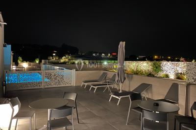Terrasse extérieure éclairée la nuit avec des tables, des chaises longues et une vue sur une piscine illuminée.