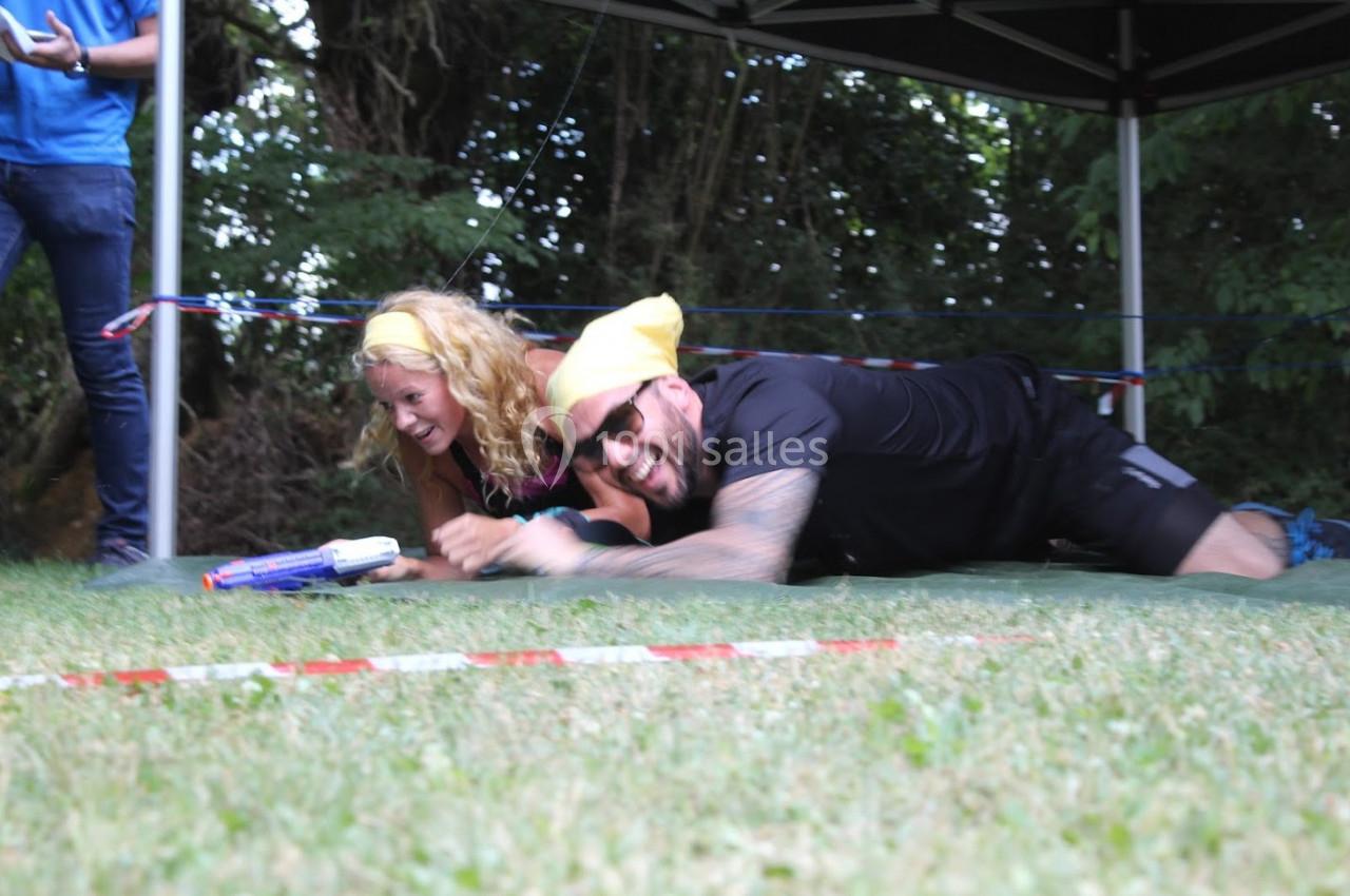 Deux personnes souriantes participent à une activité ludique en extérieur, rampant sur l'herbe sous une structure.