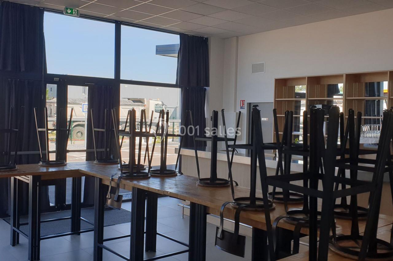 Chaises empilées sur des tables dans une salle lumineuse avec de grandes fenêtres donnant sur un parking.