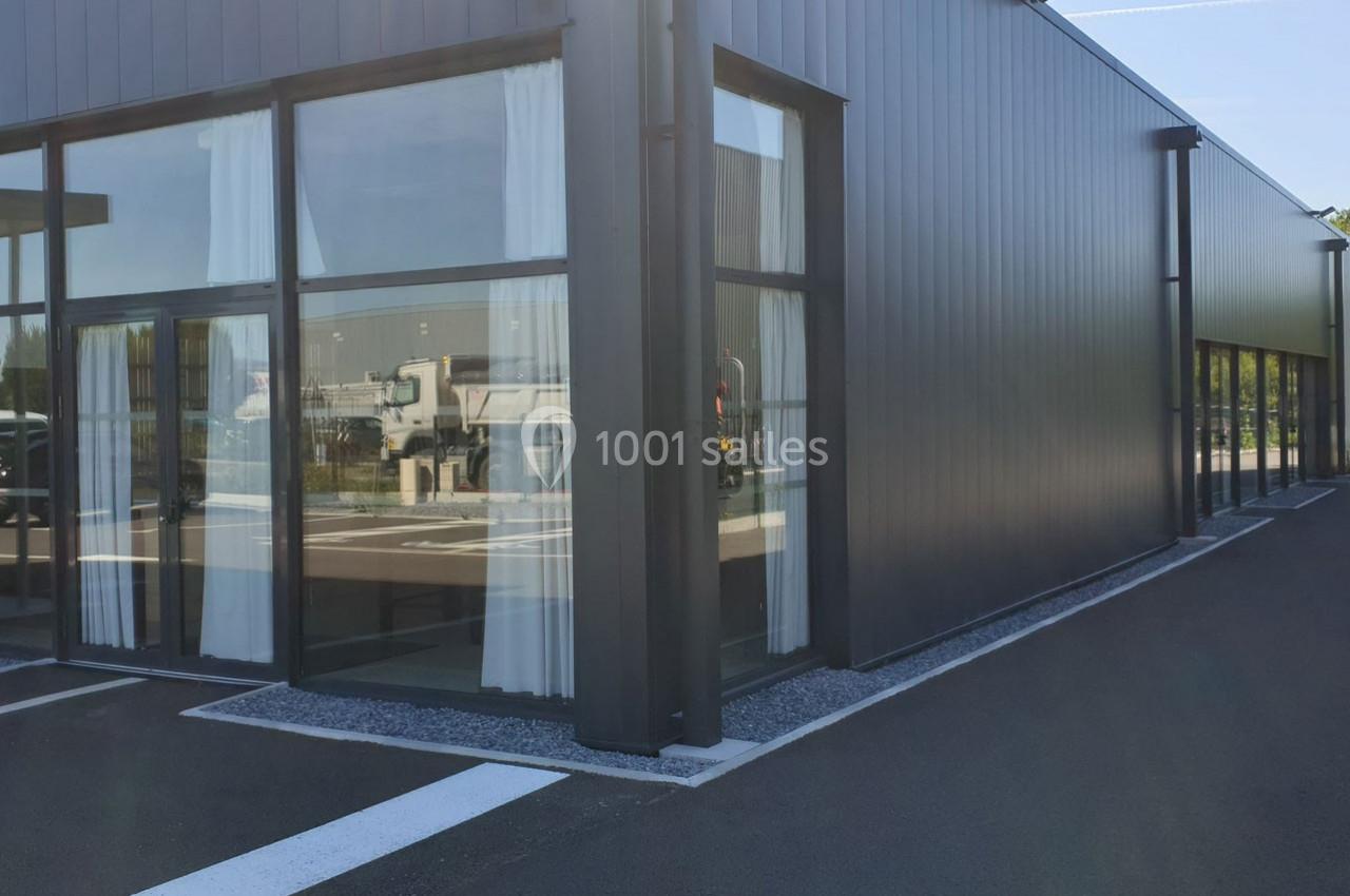 Bâtiment moderne avec façade en métal gris, grandes baies vitrées et rideaux blancs, situé près d'un parking.