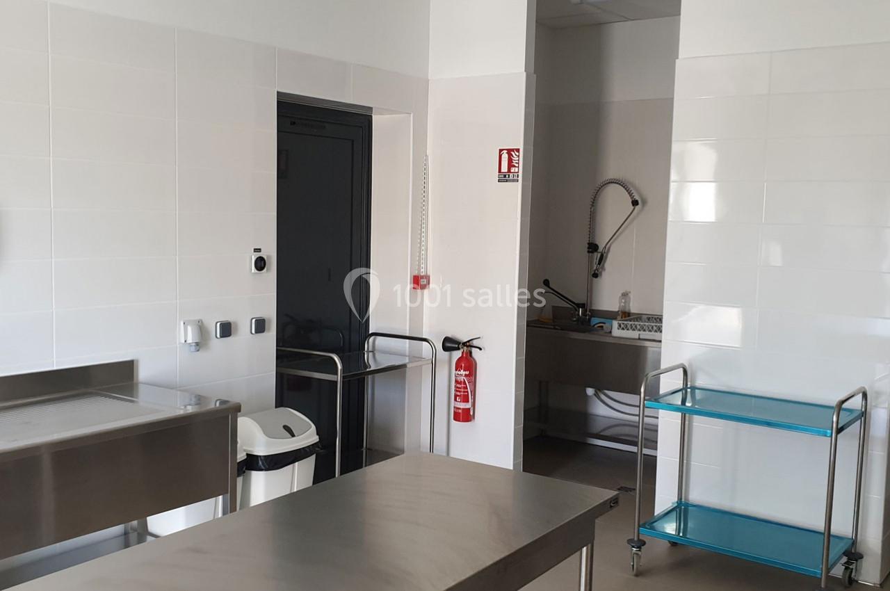 Cuisine professionnelle avec évier inox, chariot métallique, poubelle, extincteur et murs carrelés blancs.