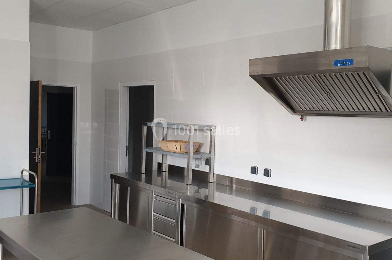 Cuisine professionnelle avec plans de travail en inox, hotte aspirante, étagère métallique et murs carrelés blancs.