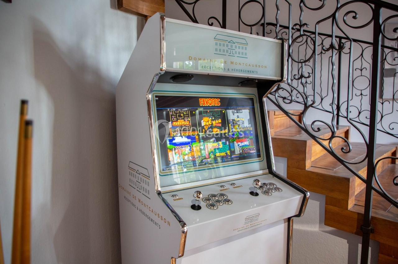 Borne d'arcade rétro blanche avec écran allumé, située près d'un escalier en bois et d'une rampe en fer forgé.