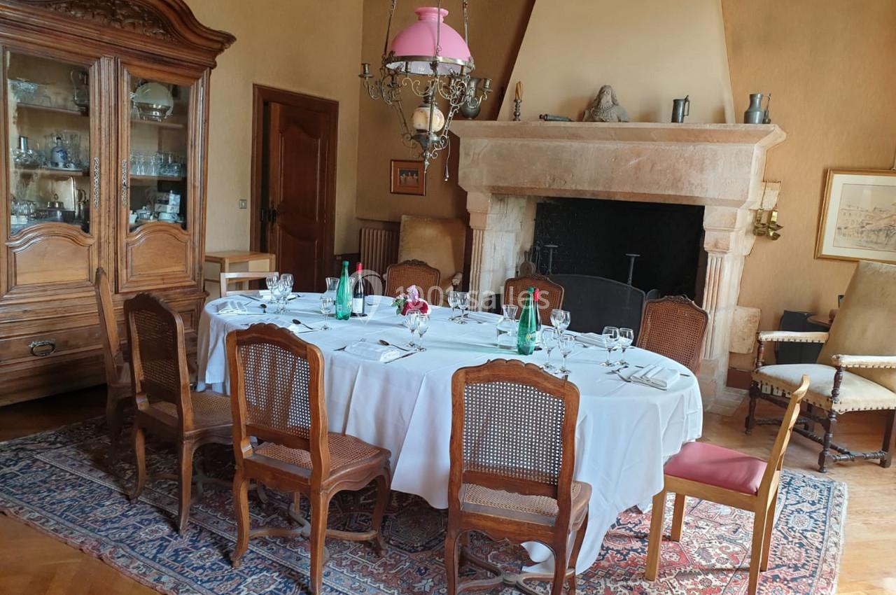 Salle à manger avec une table ronde dressée, entourée de chaises en bois, près d'une cheminée et d'un vaisselier.