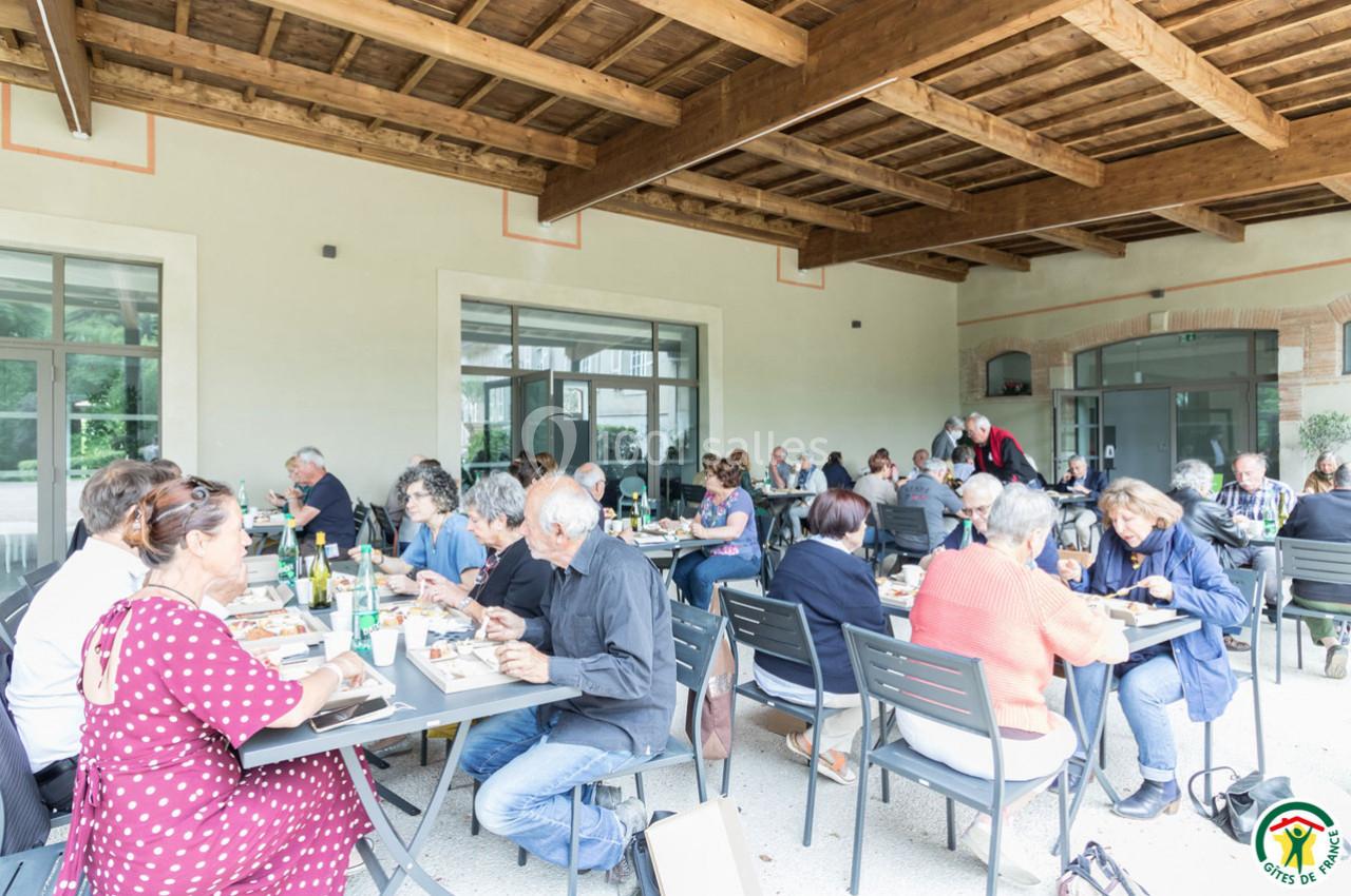Groupe de personnes partageant un repas en terrasse couverte, avec tables et chaises disposées en extérieur.