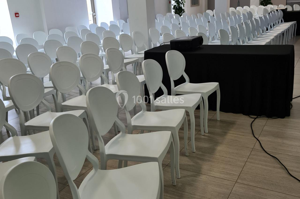 Salle de conférence lumineuse avec rangées de chaises blanches alignées et une table noire à l'avant.